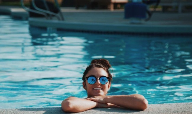 woman with sunglasses, in a pool, rests her arms on the ledge and smiles